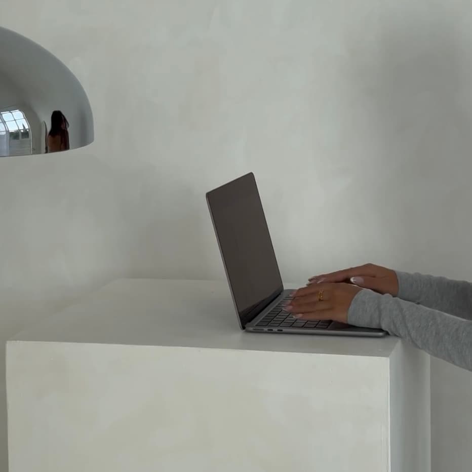 Hands in grey sleeves typing on a laptop atop a white minimalist cube.