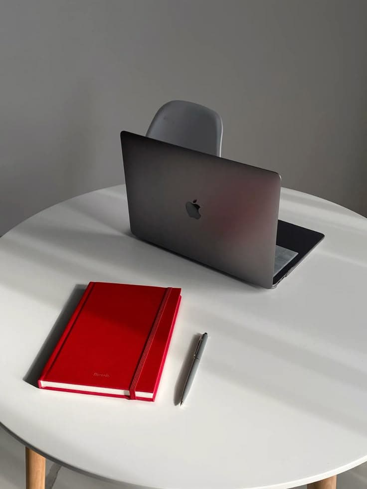 Gray laptop, red notebook, and a pen on a white table in soft natural light.